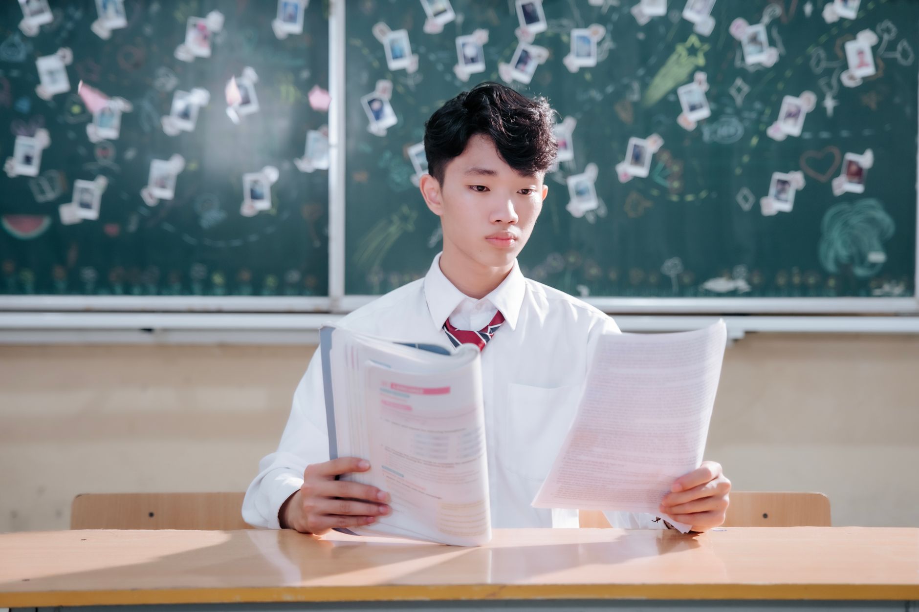 穿白衬衫系条纹领带的年轻男子坐在木桌前双手捧着一本打开的书，身后黑板上贴满白色纸片